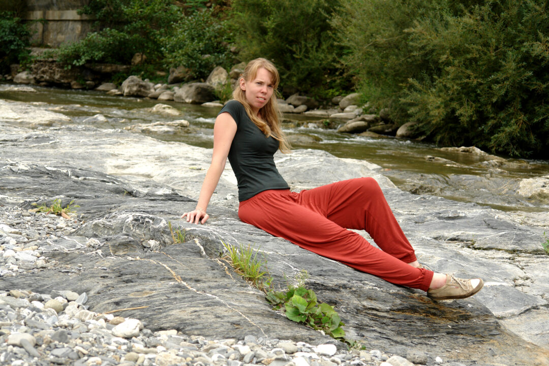 Debbie sitzt auf einem Felsen an einem Bach. Sie lehnt sich leicht zurück und stützt sich auf den Armen ab. Man sieht, dass sie sehr schlank ist.