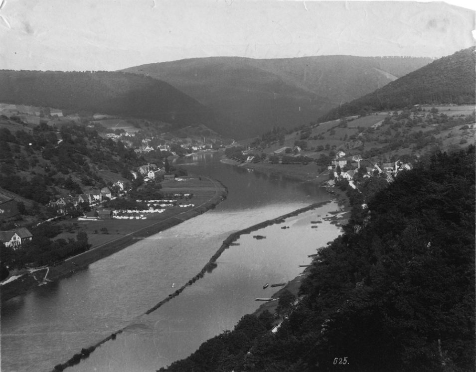 Schwarzweißfoto, das von einem erhöhten Standort zwischen Heidelberg und Ziegelhausen aufgenommen wurde. Der Blick geht Richtung Osten nach Ziegelhausen. Zu sehen ist, dass das Neckarufer viel breiter war. Zwischen Neckar und Häusern befanden sich weitläufige Wiesen, während heute dort die Bundesstraße langführt.