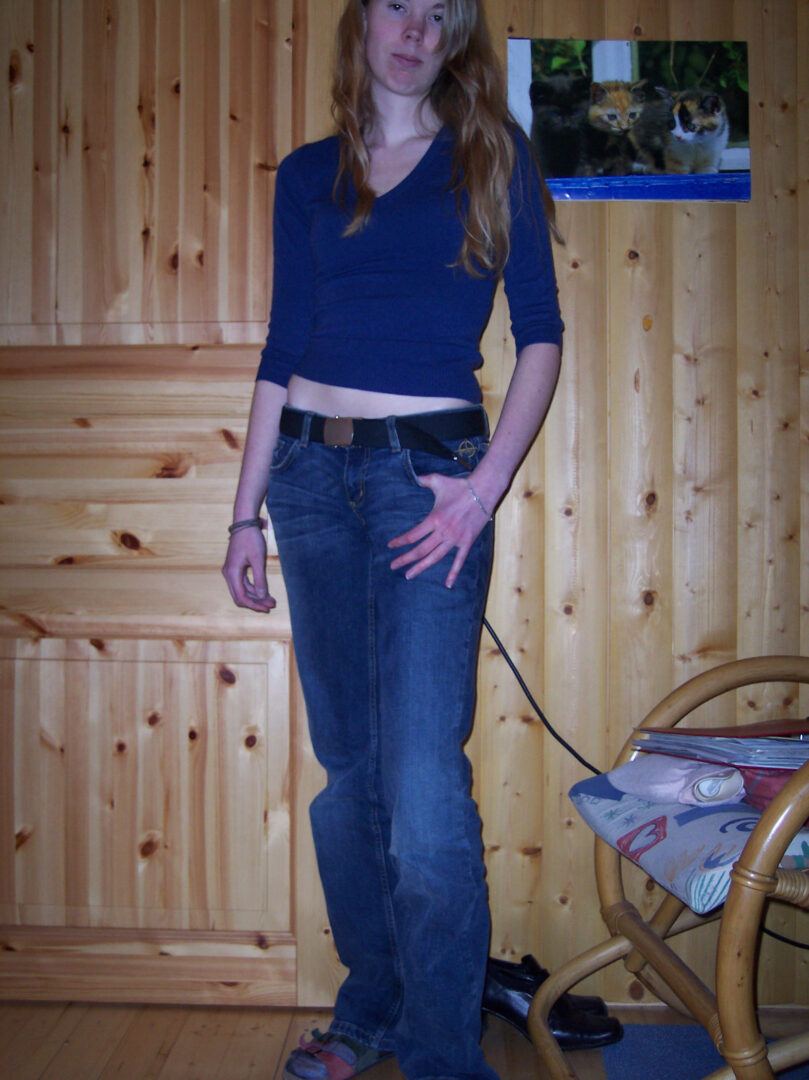 Debbie steht vor einer Holzwand und schaut in die Kamera. Das Bild dient zum Zeigen ihres Abnehmerfolgs: Sie trägt eine blaue Jeans mit Gürtel, darüber ist ein wenig Bauch zu sehen. Außerdem trägt sie einen blauen Pullover. Sie ist sehr schlank