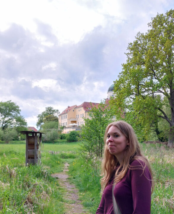 Das Foto zeigt Debbie im Vordergrund. Sie schaut verschmitzt in die Kamera. Ein schmaler Trampelpfad führt in Richtung zum beige-gelben Burggebäude im Hintergrund.