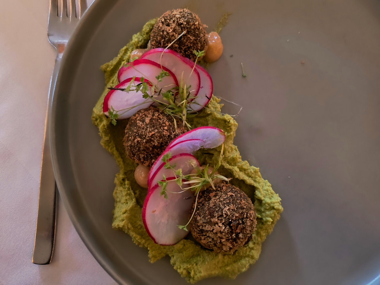 Blick auf die Pilzpralinen-Vorspeise: Falafel-artige dunkle Kugeln liegen auf grünem Erbsenpüree. Dazwischen Radieschenscheiben und frische Sprossen.