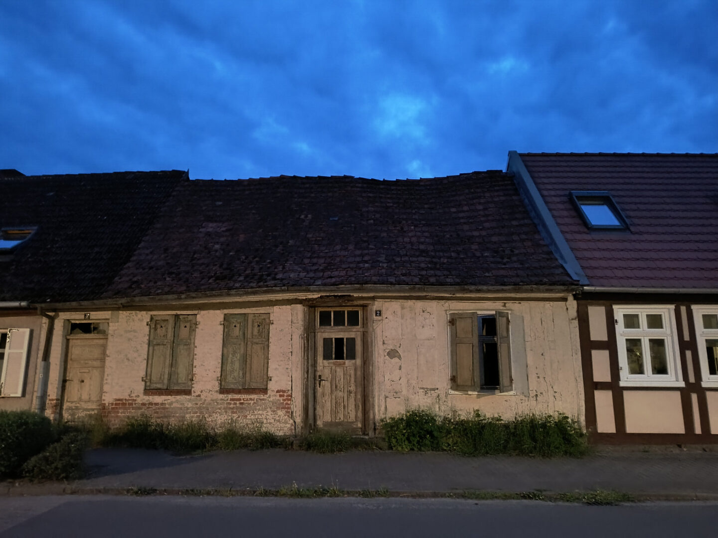 Kleine, niedrige Hütte, die direkt in ähnlich kleine Häuser links und rechts davon übergeht. Zentral ist eine Haustür, links davon zwei Fenster mit geschlossenen Fensterläden, rechts der Haustür ebenfalls ein Fenster. Die Läden sind teilweise offen. Das Haus steht leer. Auf einem schmalen Streifen zwischen Haus und Bürgersteig wächst Unkraut.