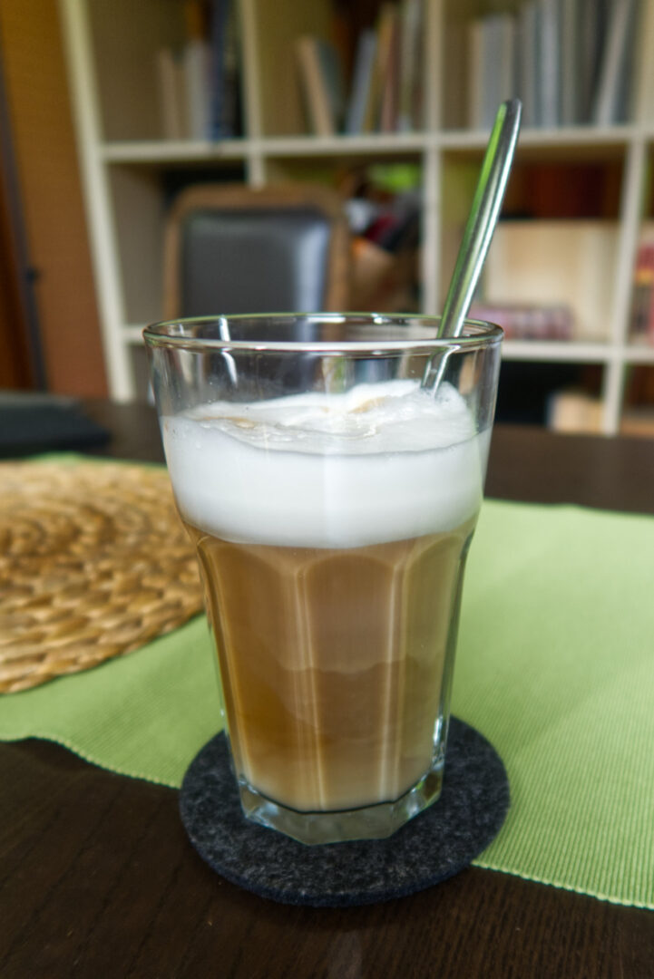 Hohes Glas mit Latte Macchiato: Oben weißer Schaum, unten der Milchkaffee. Im Hintergrund leicht verschommen mein Wohnzimmer.