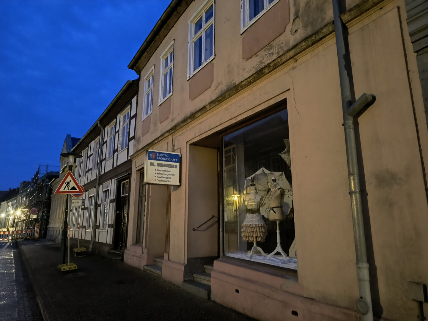 Foto eines Hauses mit Schaufenster. Im Schaufenster stehen zwei Schneiderpuppen, die aufwändige Papierkleider tragen. Das sieht eher nach Kunst aus als nach "Elektrofachgeschäft", wie auf dem Schild über dem Schaufenster zu lesen ist.