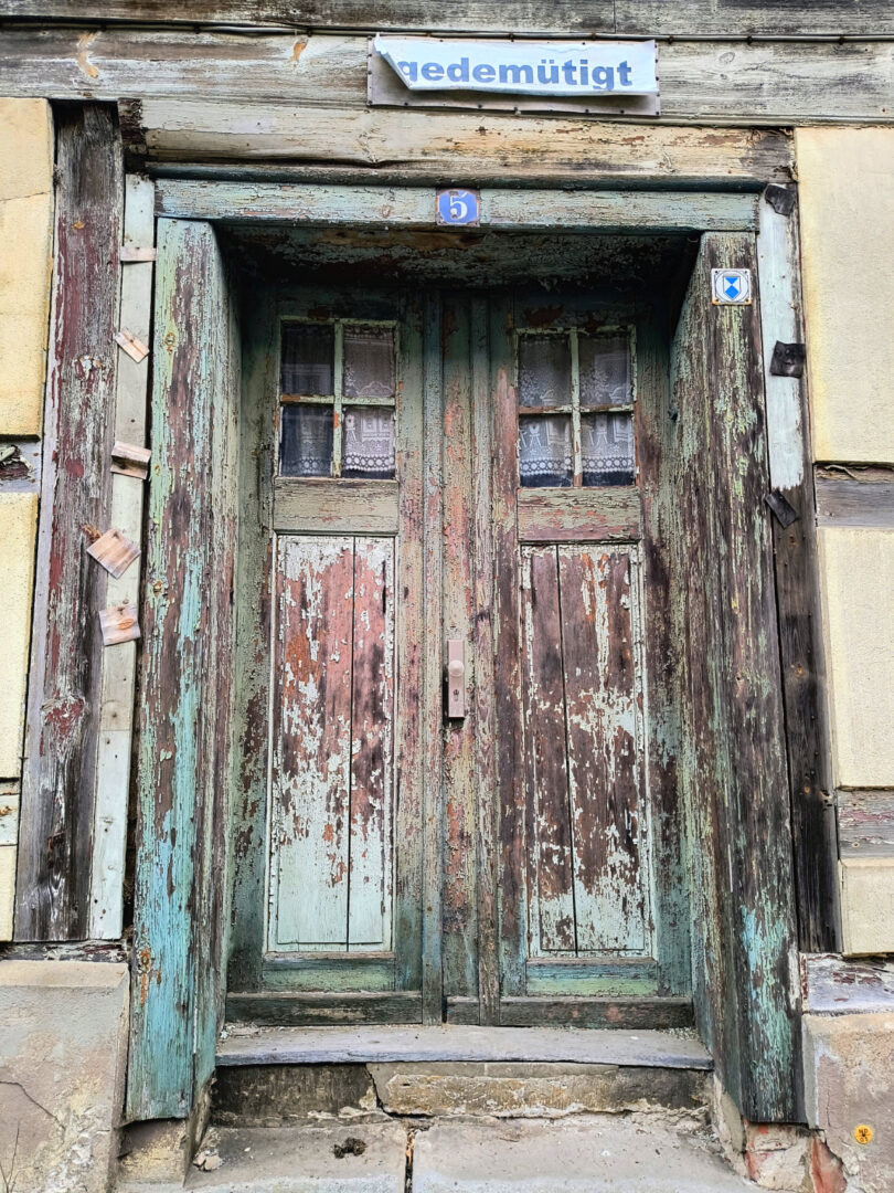 Stufen führen zu einer stark verwitterten zweiflügeligen Holz-Haustür. Im oberen Drittel der Flügel befindet sich jeweils ein Sprossenfenster, dahinter hängt eine gehäkelte, weiße Gardine. Das Haus wirkt unbewohnt.