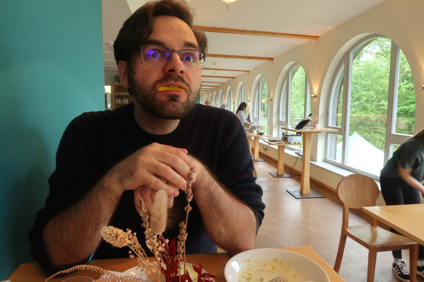 Das Foto zeigt Pierre mit einem dunklen Pulli. Er sitzt am Frühstückstisch und schaut mit einem Stück Melone im Mund an der Kamera vorbei. Er hat einen Dreitagebart und trägt eine Brille