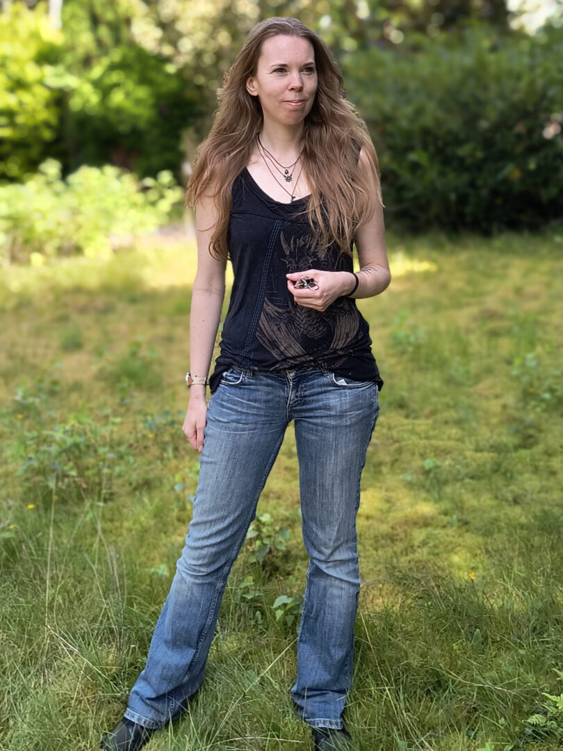 Debbie steht im Garten. Sie trägt eine blaue Jeans, ein tailliertes schwarzes ärmelloses Oberteil und um den Hals mehrere Ketten. Ihre Haare liegen aufgelöst auf den Schultern.