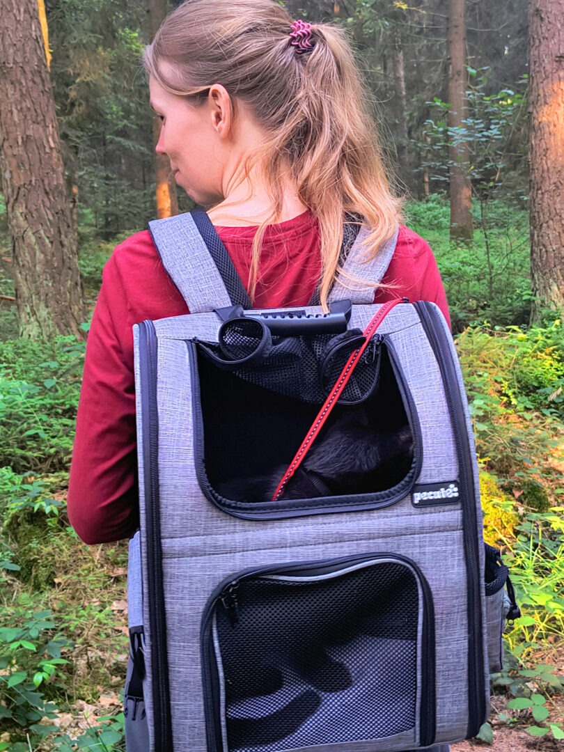 Ich stehe im Wald und trage auf meinem Rücken einen Rucksack, in dem eine schwarze Katze sitzt (Balu). An der Ober- und Rückseite befinden sich schwarze Mesh-Fenster, durch die das Tier rausschauen kann. Das obere Fenster ist aufgerollt, sodass Balu seinen Kopf rausstrecken könnte, wenn er wollte.