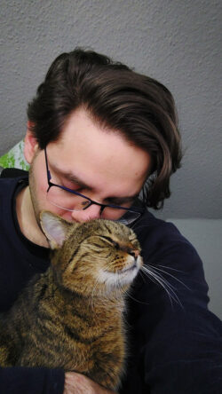 Pierre wünschte sich dieses Bild von ihm und Kater Lopi in der Galerie. Die beiden hingen sehr aneinander. Pierre, ein Mann mit braunem Haar und Brille, berührt mit seiner Nase den Hinterkopf einer getigerten Katze. Lopi hat die Augen geschlossen und scheint es zu genießen.
